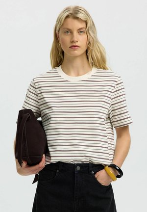 Lyshåret kvinde iført en hvid t-shirt med striber, sorte jeans, der holder en mørk taske og bærer sorte og gule armbånd på håndleddet.