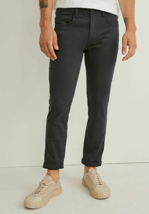 Man draagt strakke zwarte jeans met omgeslagen pijpen en beige casual sneakers, staand tegen een neutrale achtergrond.