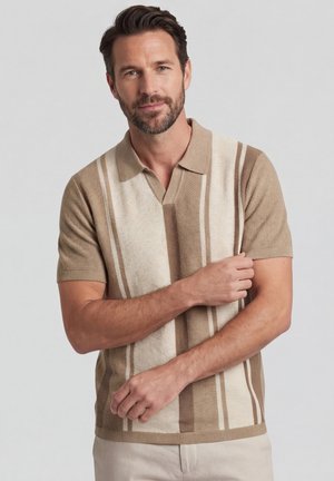 Homme aux cheveux courts et foncés et à la barbe, portant un polo en maille rayé beige et crème à manches courtes, debout devant un fond gris clair.