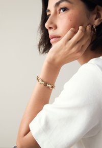 Gold metal chain bracelet with interlocking oval links worn on a wrist. The wrist is partially covered by a white short-sleeve shirt.