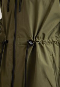 Veste vert olive avec fermeture éclair noire et cordon de serrage noir réglable à la taille montrant la texture du tissu et les détails de couture.