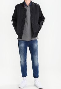 Svart bombarjacka över en grå polo, blå slitna jeans och vita sneakers. Jackan har en slät yta och ribbad mudd.