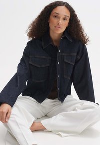 Veste en denim foncé avec fermeture à boutons, grandes poches et couture contrastante ; présente une coupe décontractée et des manches larges. Modèle assis, portant un pantalon blanc.