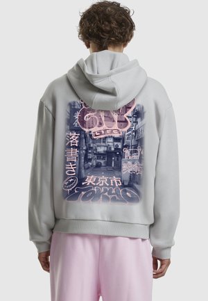 Lys grå hoodie med et stort pink grafisk design på ryggen, der viser stiliseret tekst og billeder af en bybillede. Blødt stof, afslappet pasform.