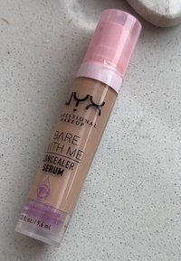 NYX Bare With Me Concealer Serum in un tubo trasparente, con un erogatore rosa e accenti viola, dotato di una formula leggera e cremosa di colore beige.