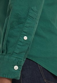 Camisa de algodón verde con manga larga, que presenta un puño abotonado y botones blancos en contraste. La tela tiene una textura suave.