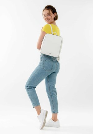 White rectangular backpack made of smooth material, featuring a top handle and shoulder straps, paired with light blue jeans and white sneakers.