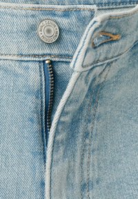 Nahaufnahme von hellblauen Jeans mit teilweise geöffnetem Reißverschluss und Metallknopf mit der Aufschrift "MNG DENIM".