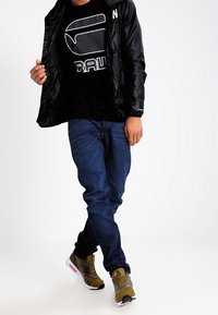 Czarna, lekka kurtka na suwak, noszona na czarnej koszulce z grafiką i białym napisem. Niebieskie dżinsy i oliwkowo-zielone sneakersy.