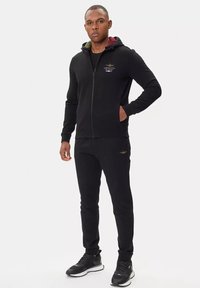 Felpa nera con zip e pantaloni jogger abbinati, con un logo sul petto, tasche e polsini a costine. Tessuto morbido con un design aderente.