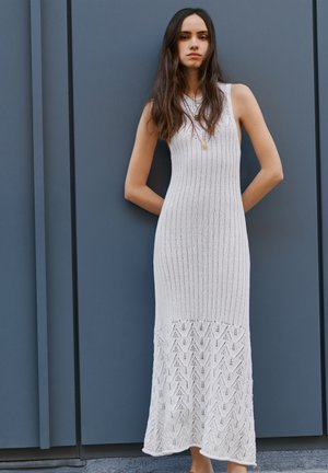 Γυναίκα με μακριά σκούρα μαλλιά που φοράει άσπρο πλεκτό μακρύ φόρεμα χωρίς μανίκια, στέκεται με τα χέρια πίσω από την πλάτη της μπροστά σε έναν γκρι τοίχο.