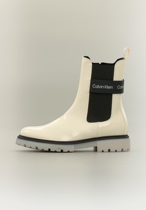 Cream-colored patent leather ankle boot with black elastic side panel and Calvin Klein logo strap on side, thick lug sole.