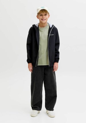 Fant, ki se smeji, nosi belo kapo, črno jakno z zadrgo čez zeleno majico, široke črne hlače in bele superge, stoji na belem ozadju.