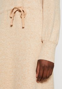 Robe-pull en tricot beige clair, avec une finition texturée, des manches longues et une ceinture en tissu à nouer à la taille pour un ajustement réglable.