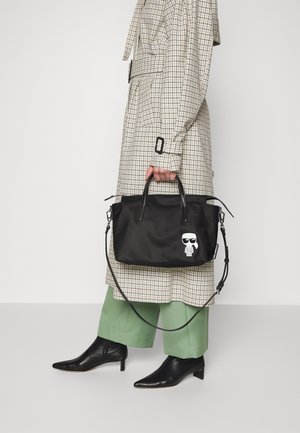 Personne portant un trench-coat beige à carreaux et un pantalon vert, tenant un sac à main noir avec une petite figure de dessin animé blanche dessus.