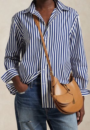 Persoon die een blauw-wit gestreept overhemd met verticale strepen en opgerolde mouwen draagt, versleten blauwe spijkerbroek en een lichtbruine leren schoudertas.