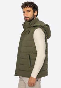 Man met donker krullend haar, gekleed in een olijfgroene gewatteerde jas met capuchon en een lichtbeige overhemd met lange mouwen, staand opzij.