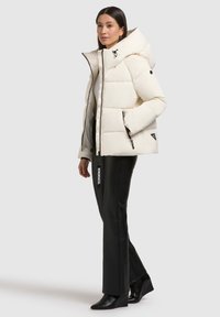 Weißer Puffer-Jacke mit hohem Kragen und Kapuze, ausgestattet mit schwarzen Reißverschlüssen und einem Logo-Tag. Getragen über schwarzen engen Hosen und Wedge-Stiefeln.