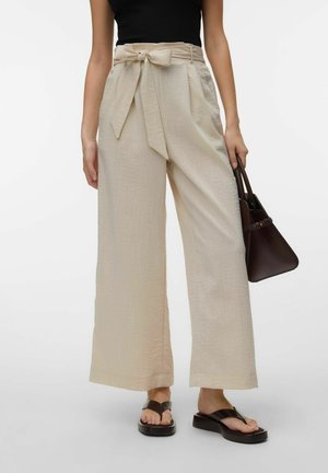 Kobieta nosząca beżowe szerokie spodnie z wiązanym paskiem, czarny top, czarne sandały na koturnie, trzymająca ciemnobrązową torebkę.