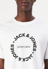 White cotton t-shirt featuring a circular black graphic with "JACK & JONES" and "DENIM CREW" text centered in the front. Short sleeves.