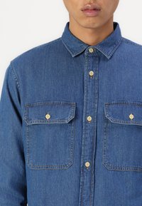 Denim shirt in medium blue, featuring a collar, two chest pockets with button closures, and contrasting yellow stitching.