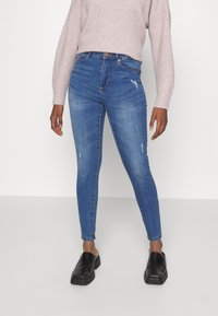 Kvinna i blå högmidjade skinny jeans med mindre slitage, ljusrosa tröja och svarta slip-on skor, stående mot en neutral bakgrund.