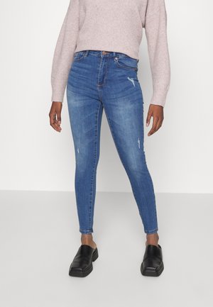 Kvinna i blå högmidjade skinny jeans med mindre slitage, ljusrosa tröja och svarta slip-on skor, stående mot en neutral bakgrund.