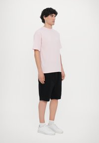 Rosa Baumwoll-T-Shirt mit kurzen Ärmeln, kombiniert mit schwarzen Shorts. Das Modell trägt weiße Turnschuhe und steht vor einem schlichten weißen Hintergrund.