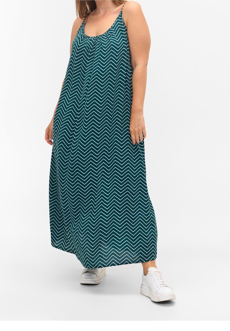 Robe maxi en teal avec un motif chevron blanc, design sans manches, tissu fluide et encolure froncée, portée avec des baskets blanches.