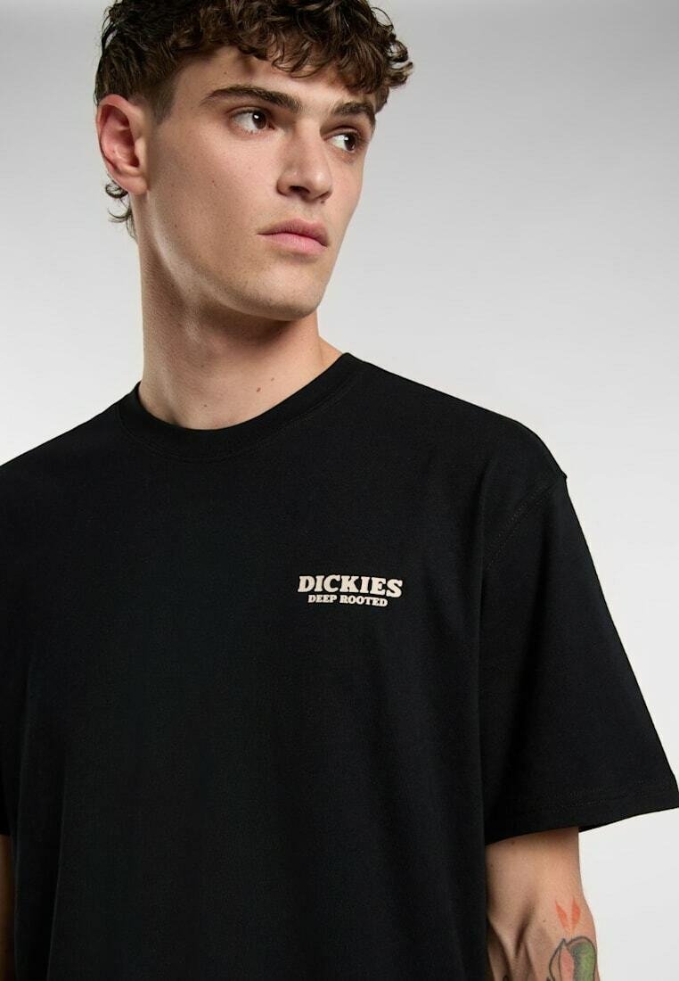 Ung mand med krøllet hår iført en sort Dickies T-shirt med teksten "Deep Rooted", der kigger til siden mod en ensartet lys baggrund.