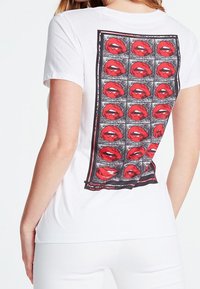 Camiseta de algodón blanca con un gran estampado gráfico de labios rojos sobre un fondo de cuadrícula negra, con texto cerca de la parte inferior. Mangas cortas y ajuste relajado.