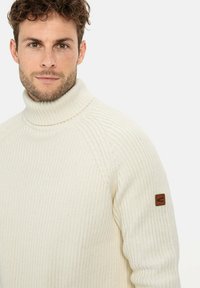 Cremfarbener Rollkragenpullover mit geripptem Muster, ausgestattet mit einem Lederlogo-Patch auf dem linken Ärmel und einer taillierten Passform.
