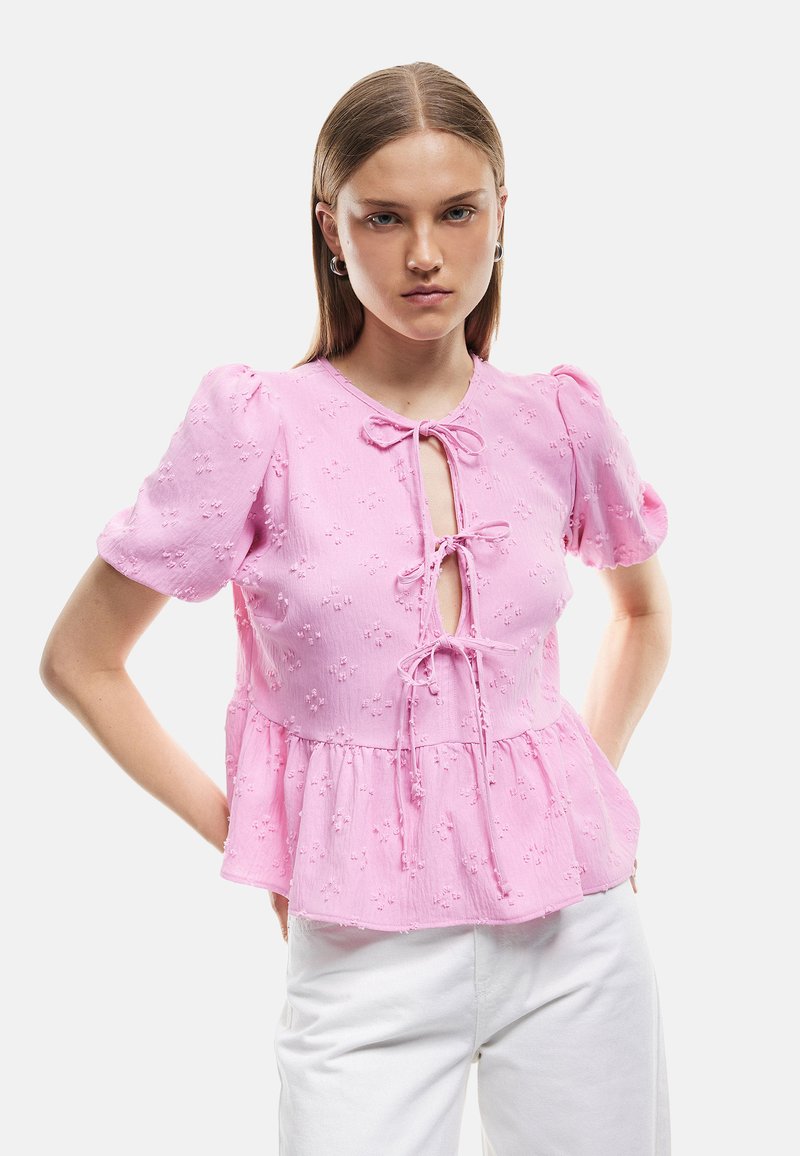 Vaaleanpunainen peplum-toppi, jossa on lyhyet puff-hihat, edessä solmittava suljenta, kukkakirjontaa ja teksturoitu kangas. Yhdistettynä valkoisiin housuihin.