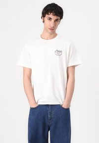 Weißes Baumwoll-T-Shirt mit einem kleinen schwarzen Grafikdesign auf der linken Brust. Rundhalsausschnitt, kurze Ärmel und lockerer Schnitt. Getragen mit blauen Jeans.