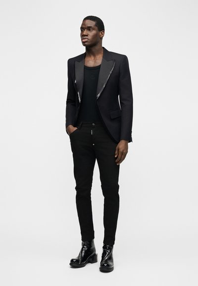 Νεαρός άνδρας στέκεται φορώντας ένα μαύρο σακάκι με σατέν πέτο, μαύρη φανέλα, στενά μαύρα παντελόνια και γυαλιστερά μαύρα μποτάκια.