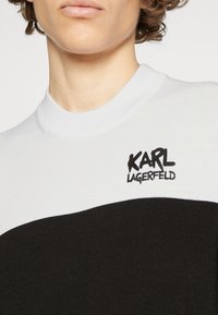 Sweter z dzianiny w białym i czarnym kolorze z wysokim kołnierzem. Zawiera logo "KARL LAGERFELD" w kolorze czarnym. Gładka struktura i dopasowany krój.