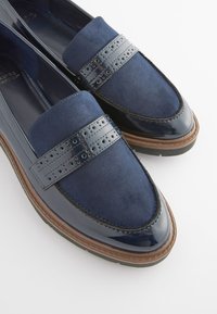 Mocassins bleu marine avec mélange de daim et de cuir verni, détails brogue sur la bride, et semelles en caoutchouc marron, pour un style décontracté ou formel.