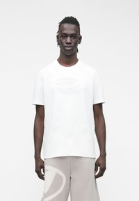 Jaunas vyras su trumpais dreadlockais, dėvintis baltą marškinėlį su reljefiniu logotipu ir šviesiai pilkus šortus su dideliu logotipu kairėje kojos pusėje.