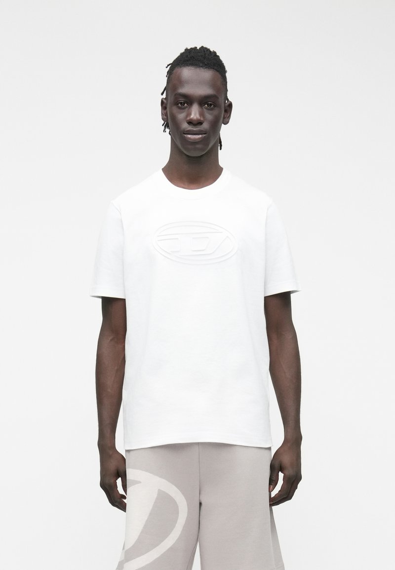 Jaunas vyras su trumpais dreadlockais, dėvintis baltą marškinėlį su reljefiniu logotipu ir šviesiai pilkus šortus su dideliu logotipu kairėje kojos pusėje.