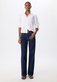 Chemise blanche à manches longues, boutonnière et manches retroussées, associée à un jean évasé bleu foncé avec des coutures contrastantes et des poches avant.