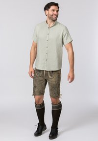 Hellgrünes, kurzärmliges Hemd mit Knöpfen, kombiniert mit braunen Leder-Shorts mit floralem Stickmuster; getragen mit dunklen, knielangen Socken und schwarzen Schuhen.