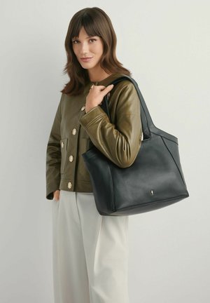 Kobieta ubraną w oliwkowo-zieloną kurtkę i białe spodnie, trzymająca dużą czarną skórzaną torbę na ramię na jednolitym tle.