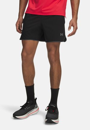 Mann trägt schwarze Sportshorts, schwarze Kniestrümpfe und schwarze Laufschuhe mit weißen Sohlen, steht vor einem einfarbigen Hintergrund.