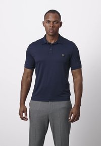 Granatowa koszulka polo z krótkim rękawem, z kołnierzykiem i małym trójkątnym logo na piersi. Noszona z szarymi spodniami.