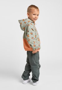 Veste légère à capuche en tissu vert avec des motifs cartoon orange, associée à un pantalon gris élastique et des baskets blanches.