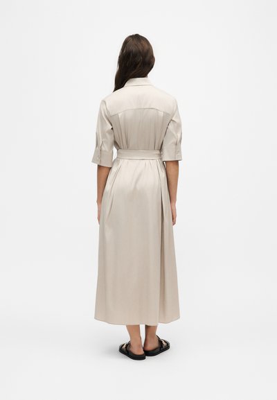 Femme portant une robe midi beige cintrée avec des manches aux coudes et des sandales noires à bout ouvert, debout de dos sur un fond blanc.