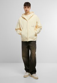 Gul zip-up hoodie med en afslappet pasform, kængurulommer og broderet tekst. Parret med mørke jeans og hvide sneakers.