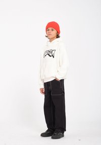 Witte hoodie met een zwart grafisch logo, gecombineerd met zwarte wijde broek met contrasterende witte stiksels, en zwarte sneakers.