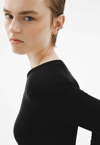 Camiseta negra de manga larga con un diseño ajustado. El modelo presenta un peinado corto y slicked-back, y lleva múltiples pendientes, incluyendo un acento azul.