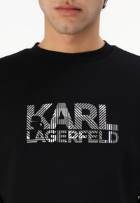 Fekete pulóver nagy, fényes ezüst grafikával, amely váltakozó csíkokban formálja a "KARL LAGERFELD" feliratot. Kerek nyakú kialakítás.